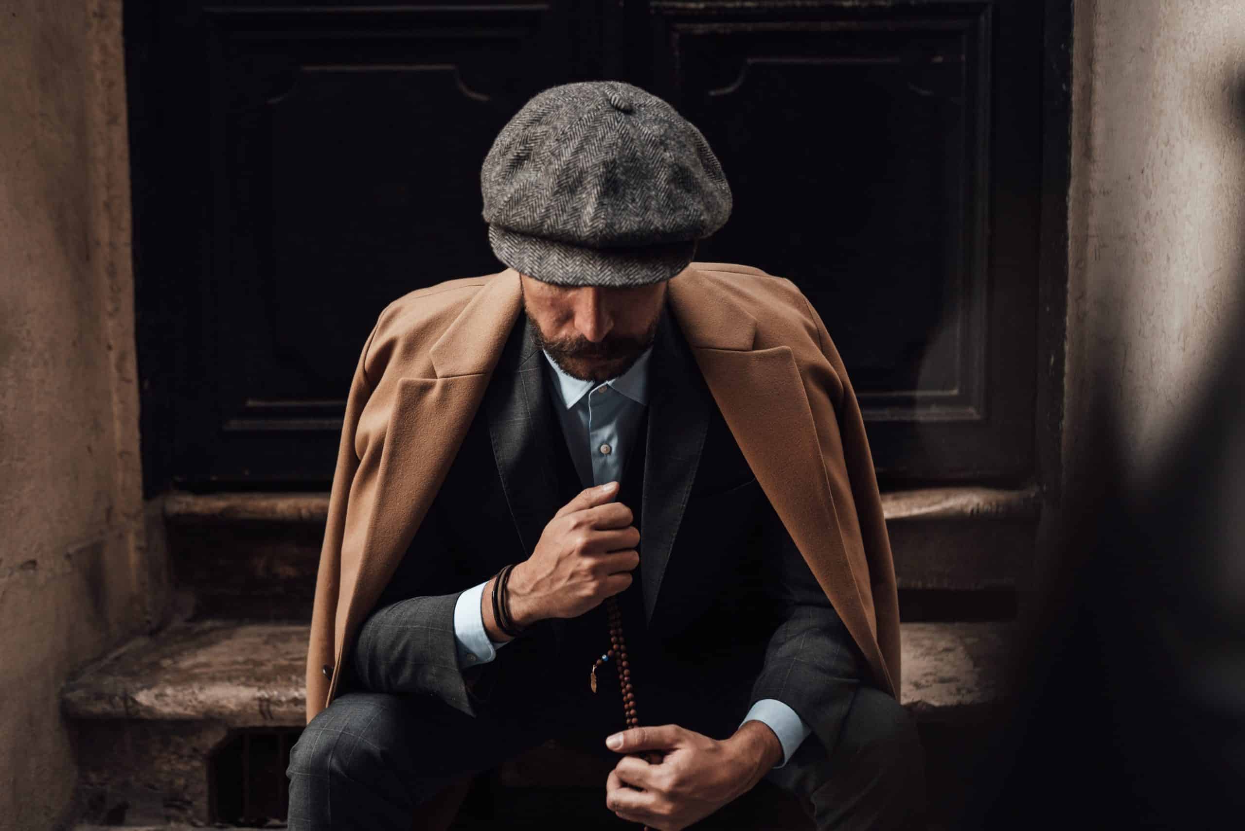 Modern gentleman seated on steps wearing a tailored camel overcoat, grey wool trousers, and a herringbone flat cap, embodying sophisticated winter style.