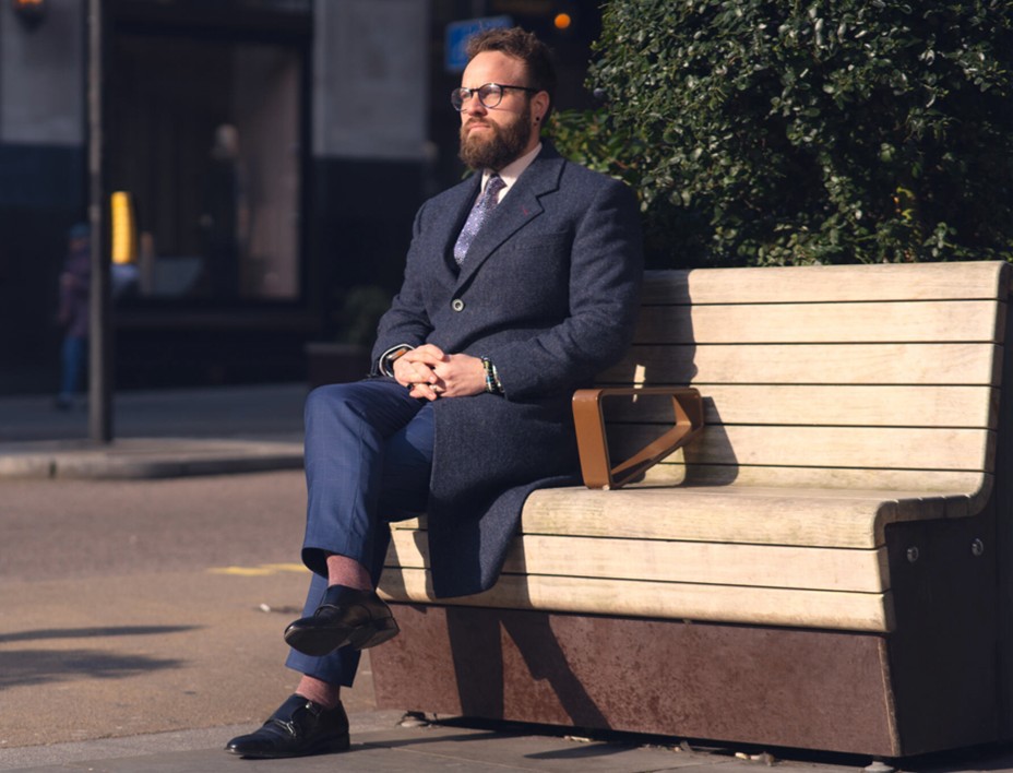 Man in blue trousers and check blazer sitting on a bench, smart with a relaxed twist.