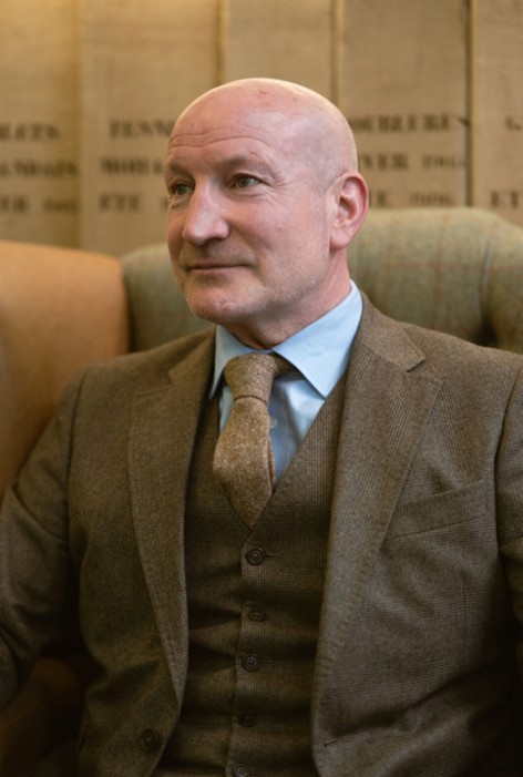 Man in a brown three-piece suit with a blue tie, classic British tailoring indoors.