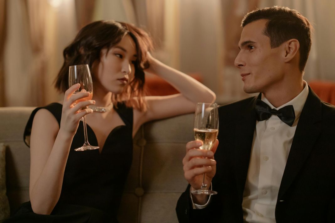 a photo of a man wearing black tie attire drinking champagne with a female guest who is wearing a black dress