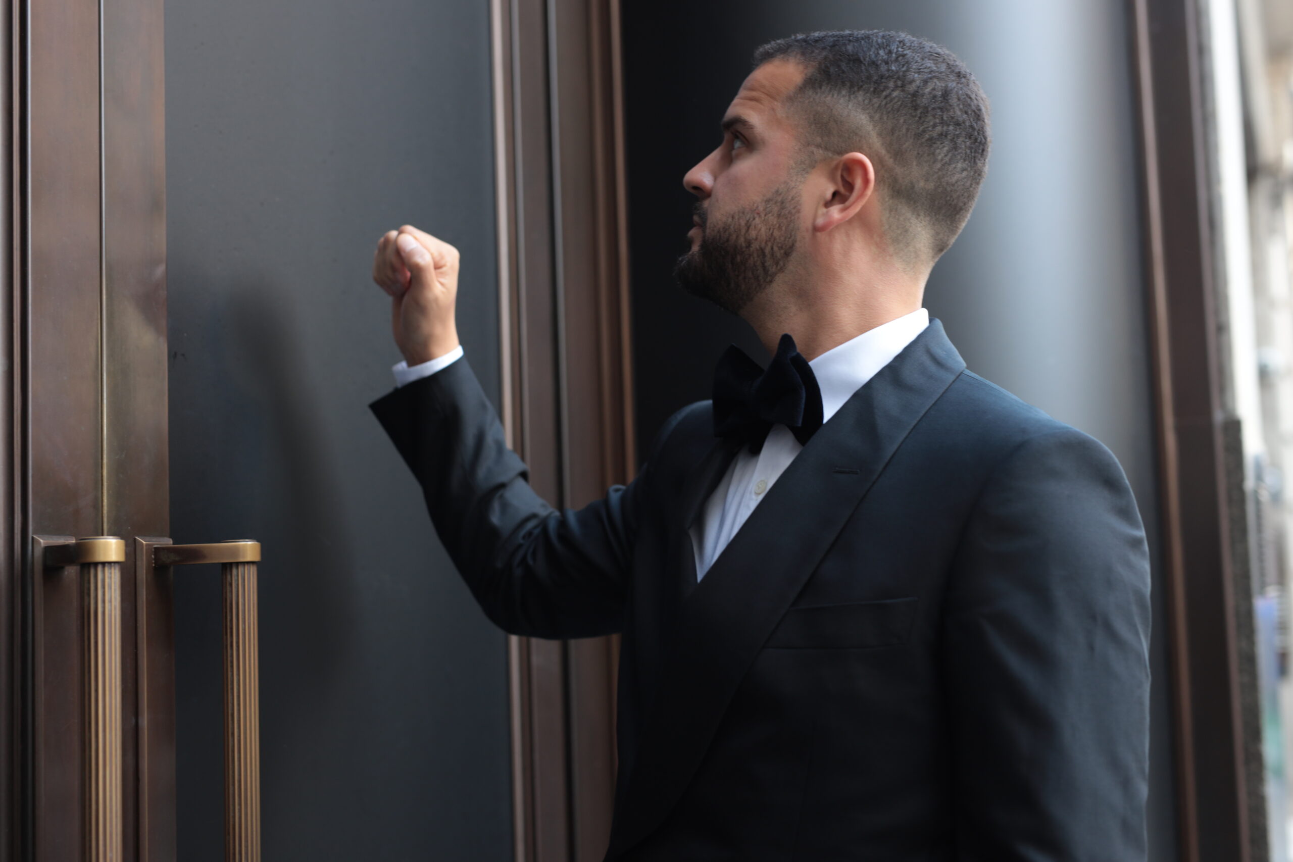 A photo of A gentleman dressed in black tie formal attire knocking on the entrance to the private black tie event