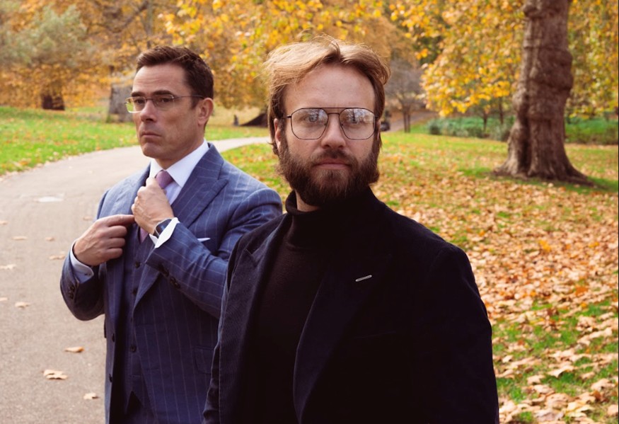 Two men wearing tailored suits in a London park during autumn, showcasing classic British menswear style.