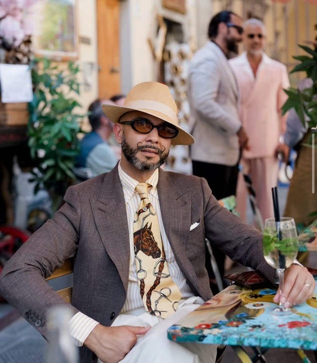 A relaxed summer tailoring look featuring a brown blazer, patterned shirt, and Panama-style hat in an outdoor setting.