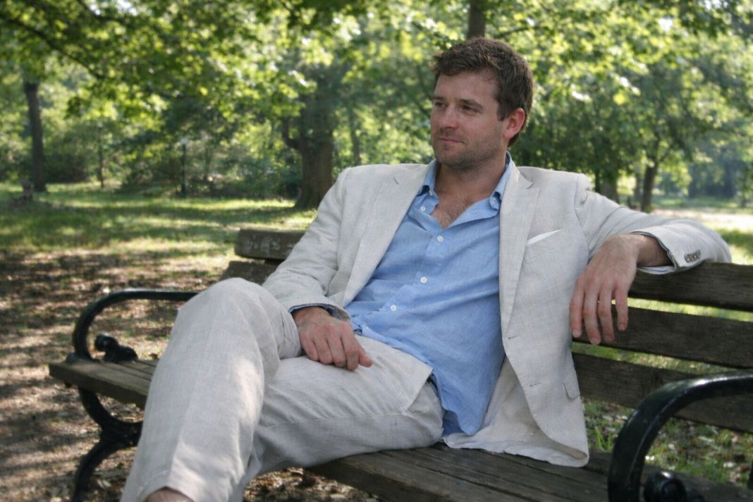An illustrative image of a man in a linen suit sitting on a park bench during a cool May morning