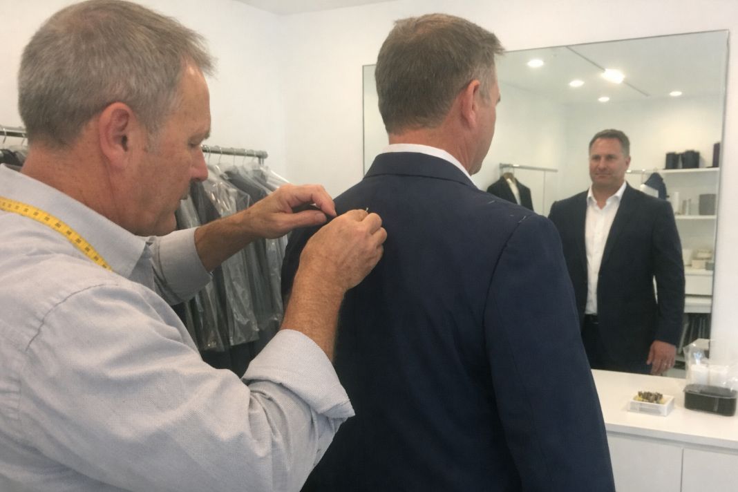 an illustrative photo of a client standing in front of a mirror while a tailor adjusts the shoulder seam during a fitting