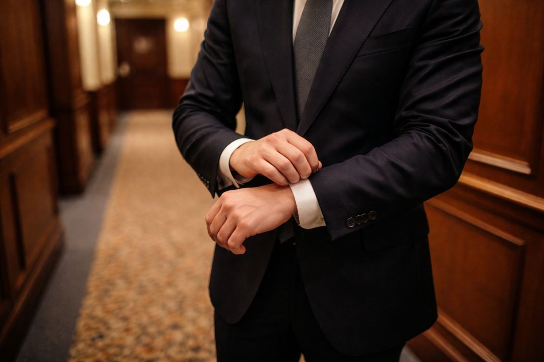 an illustrative photo of a person adjusting the cuffs of a bespoke dark navy suit in a hallway with formal carpet and wood panelling