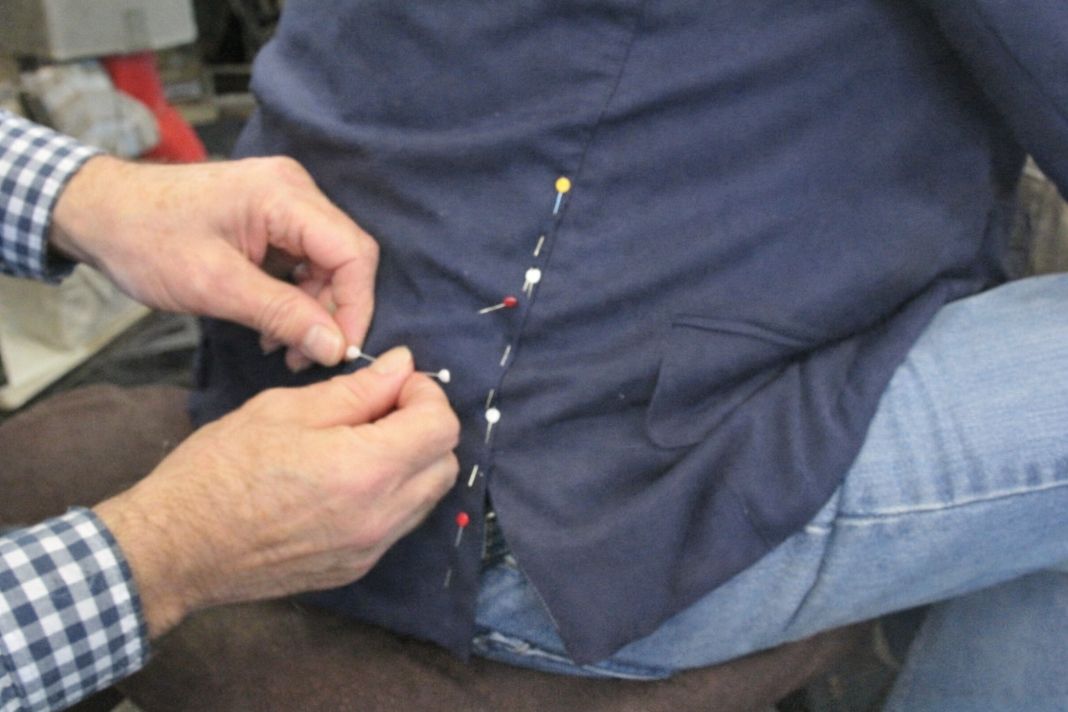An illustrative photo of a close-up view of a tailor pinning the side seam of a navy jacket worn by a seated client.