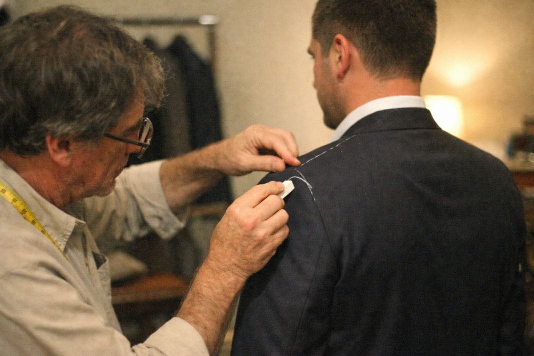 An illustrative photo of a tailor gently marking a man’s jacket near the shoulder during a fitting in a softly lit private showroom