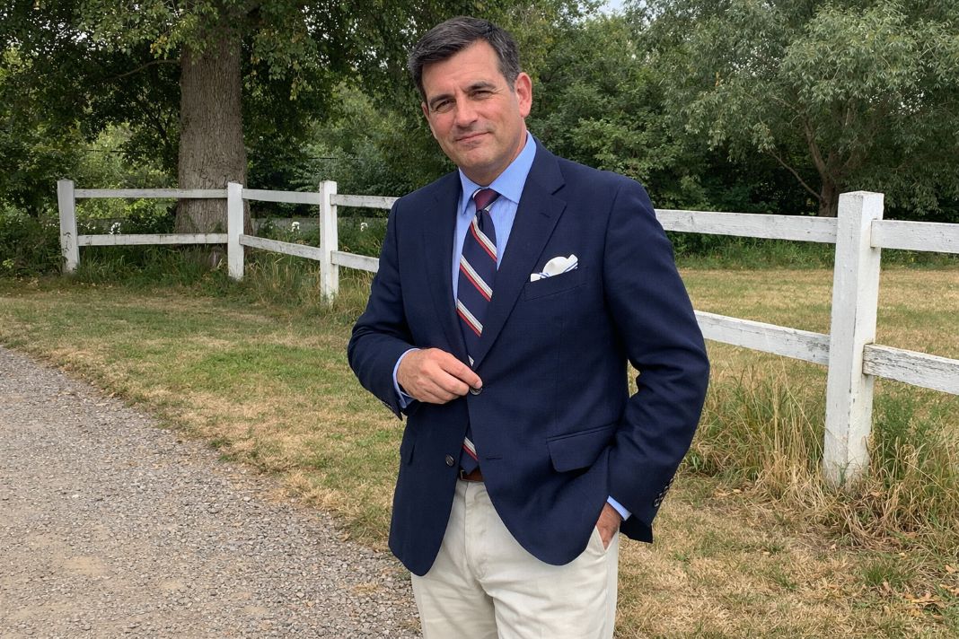An illustrative image of a cut navy blazer, pale blue shirt, repp-style tie, cream tailored trousers, brown leather loafers