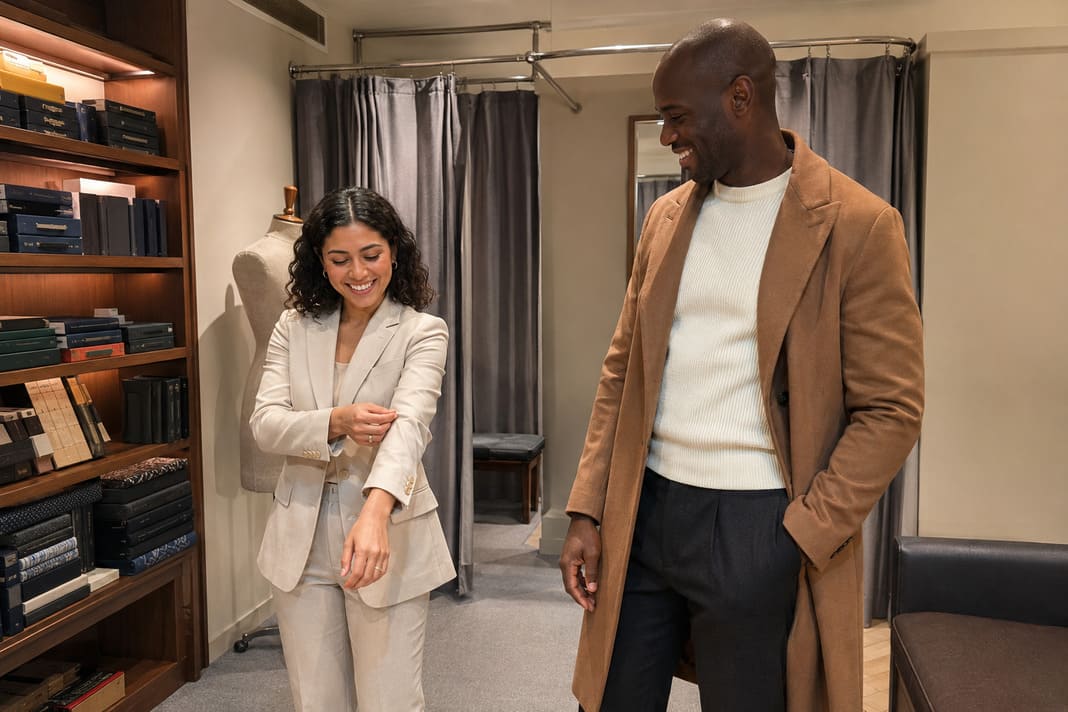 A camel blazer with dark tailored trousers creates a relaxed smart-casual look for the man, while the woman wears an ivory trouser suit with soft tailoring, demonstrating bespoke clothing designed for different body shapes, personal styles and fit preferences.