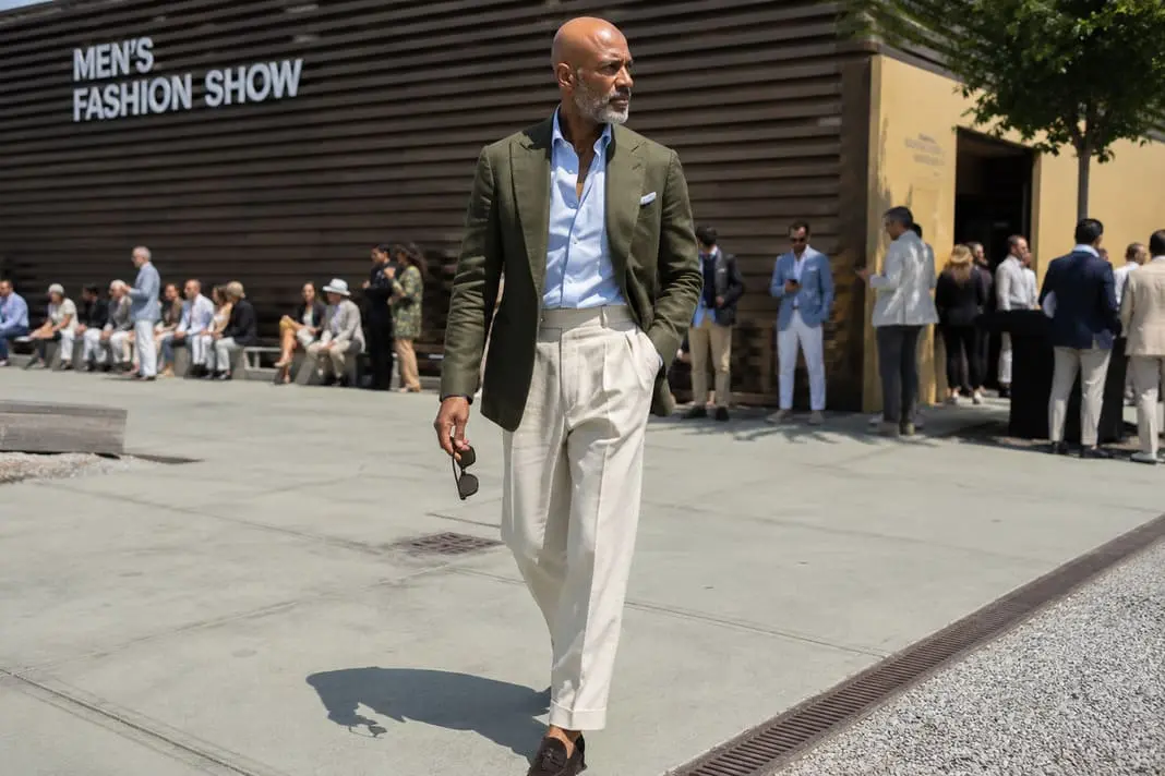 Man wearing an olive green soft-shouldered blazer with a pale blue open-neck shirt, cream pleated trousers and dark brown suede loafers, showcasing modern Pitti Uomo tailoring.