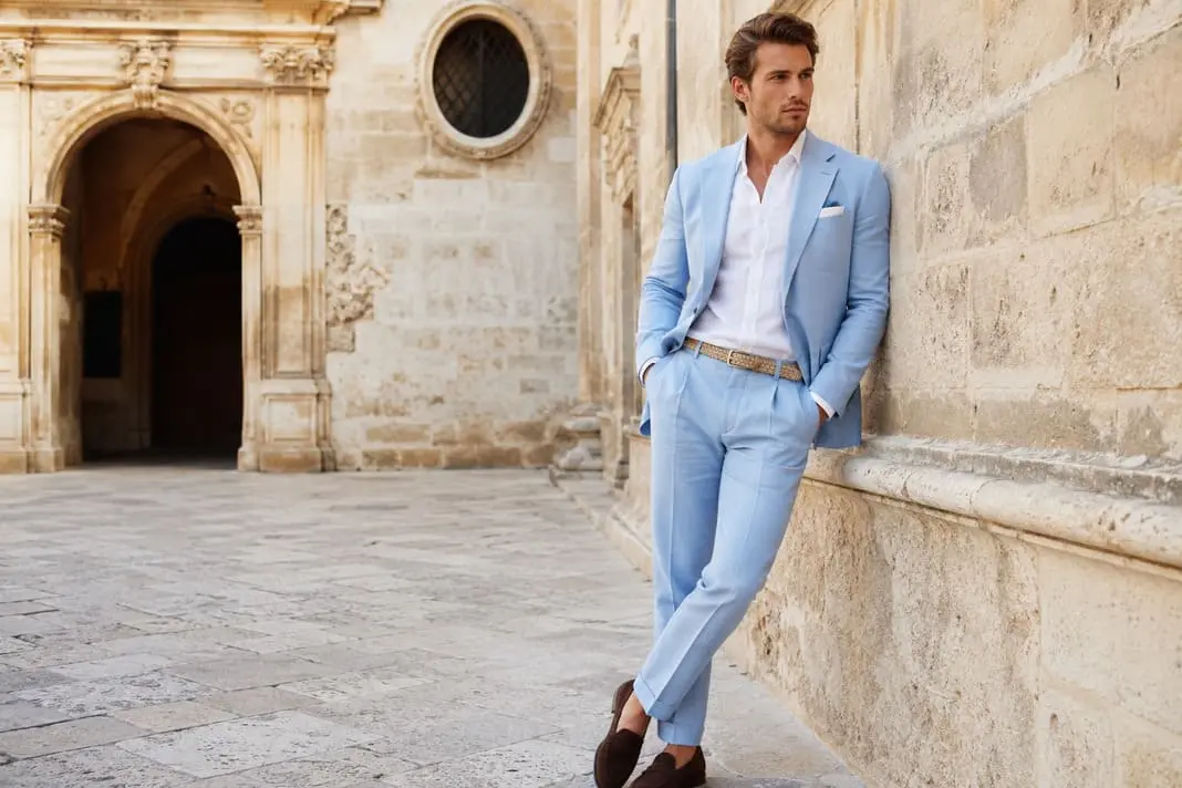 Man wearing a light blue linen suit with a white open-collar shirt, woven tan belt, patterned pocket square and brown suede tassel loafers for refined Pitti Uomo summer tailoring.