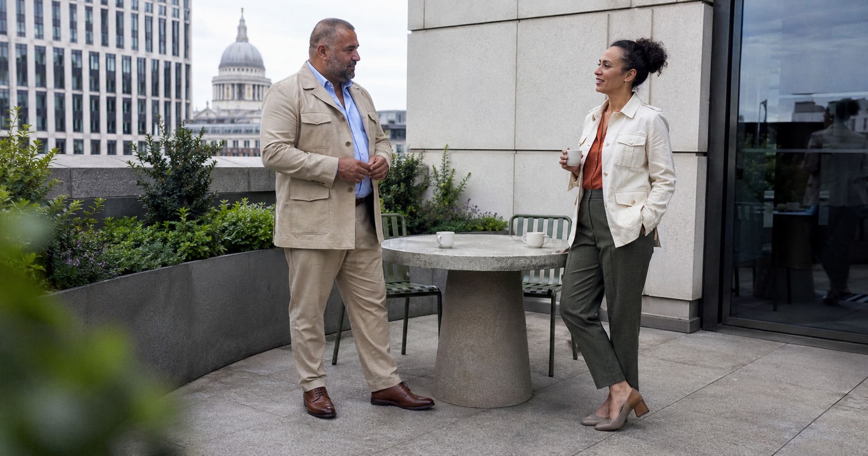 Man wearing safari jacket and woman wearing rust tailoring discuss modern safari suit styling. Man wearing safari jacket and woman wearing rust tailoring discuss modern safari suit styling.