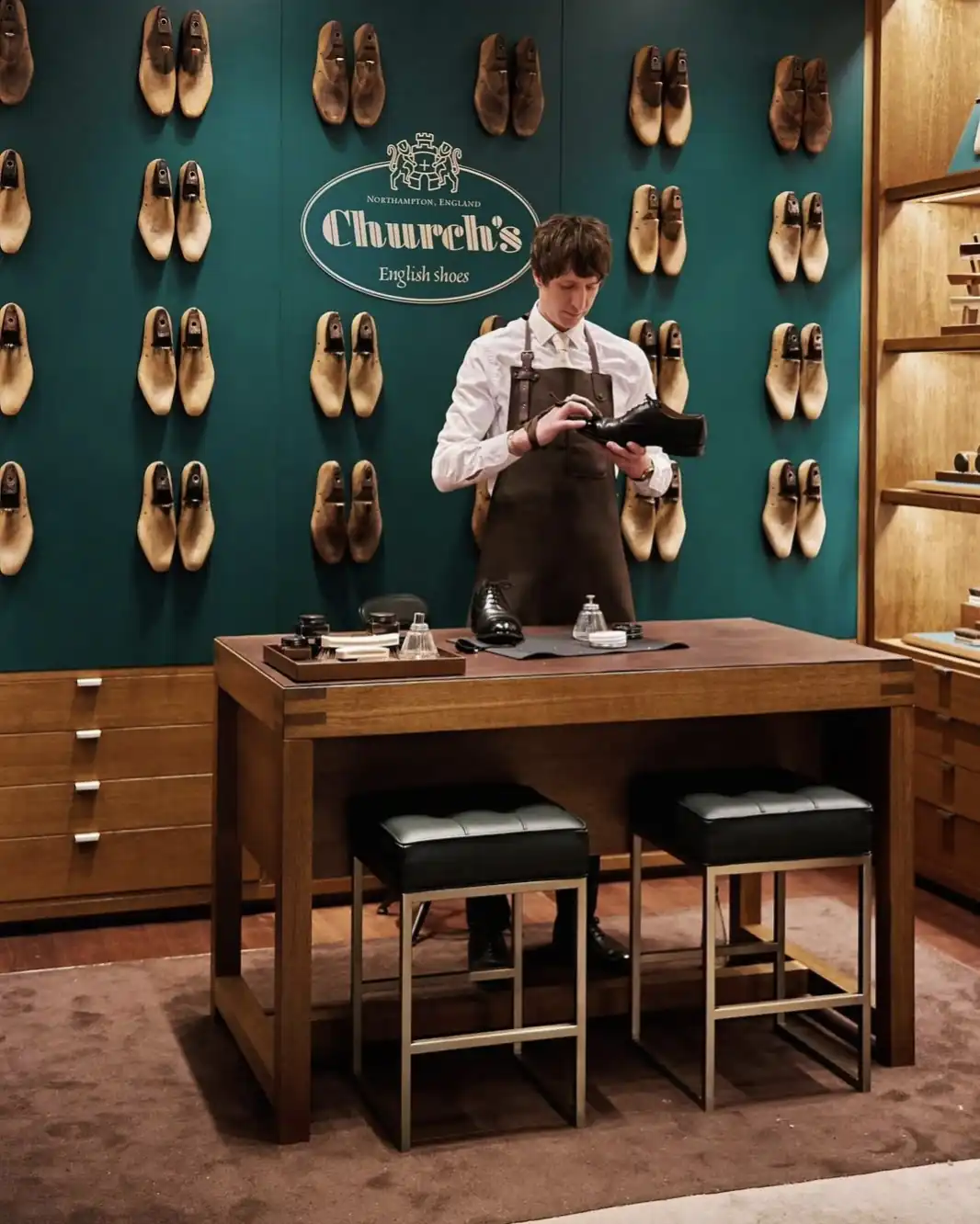 Shoe specialist in a leather apron polishing a black shoe beside a wall of lasts